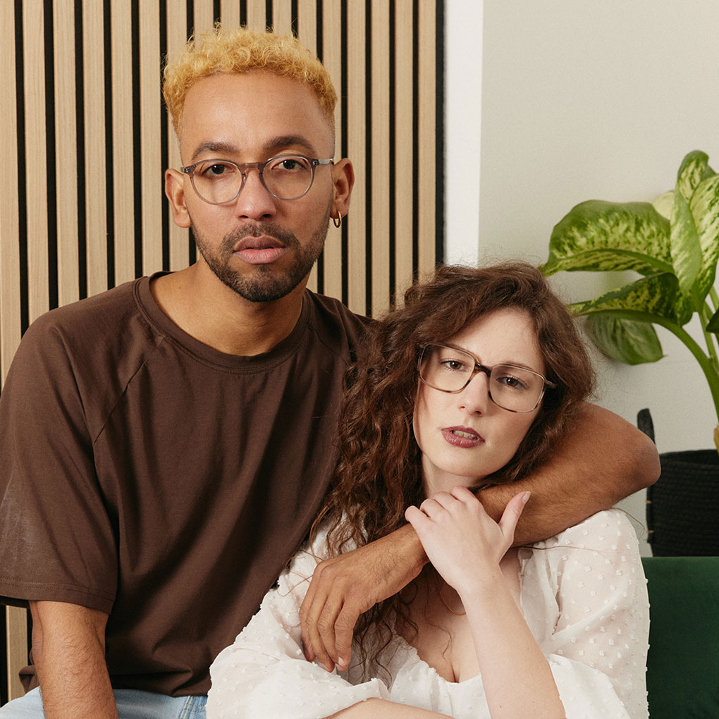 Bei FRAIMS kannst du deine Brille 10 Tage lang testen Pärchen sitzt auf dem Sofa. Sie probieren die Brillen FRAIMS Nature Tallis und Riley auf