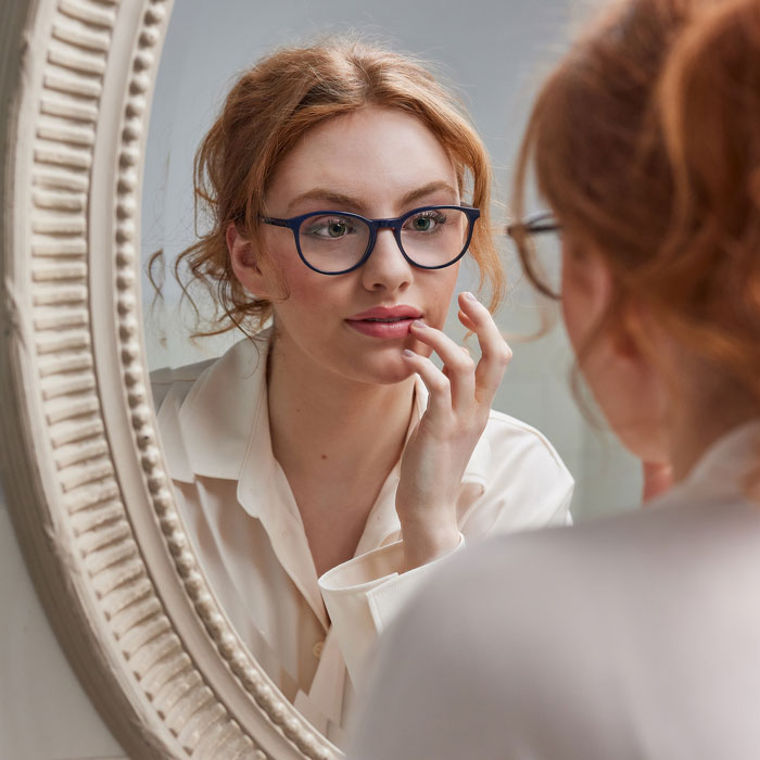 Bei FRAIMS kannst du deine Brille 10 Tage lang testen rothaarige Frau schaut in einen Spiegel. Sie trägt die Brille FRAIMS Nature Viola